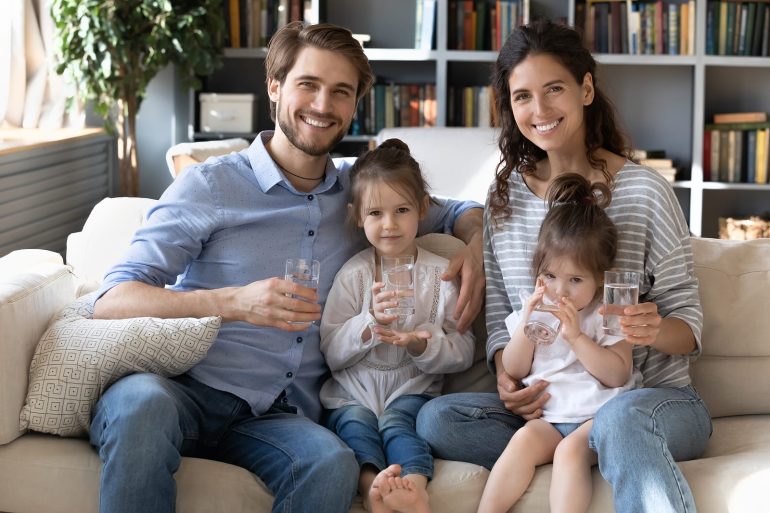 A happy family of four—parents and two young daughters—sitting together on a couch, smiling and holding glasses of water. The image promotes the importance of clean drinking water and family wellness.