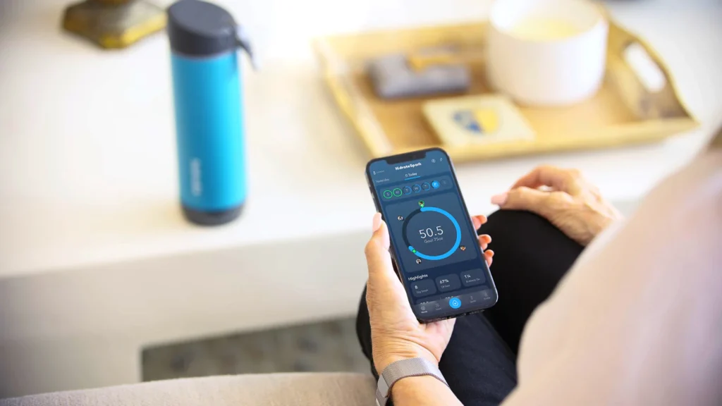 A person using a hydration tracking app on a smartphone, showing water intake data synced with a smart bottle, with a blue On Take Water bottle placed nearby on a table — promoting smart hydration and tech-enabled water tracking.

