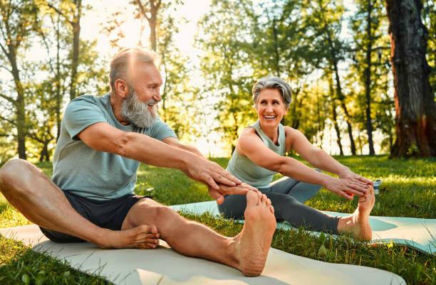 Senior couple doing outdoor yoga and stretching on mats in a park, smiling and staying active in the morning sunlight. Promotes healthy lifestyle, hydration, and wellness. Perfect representation of fitness, aging well, and staying hydrated with On Take Water.


