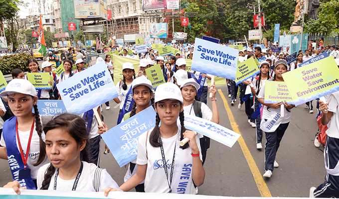 A large group of schoolchildren march in a street rally holding signs that read “Save Water Save Life” and “Save Water Save Environment.” The students are dressed in white and blue uniforms, wearing caps, and passionately promoting water conservation and awareness.