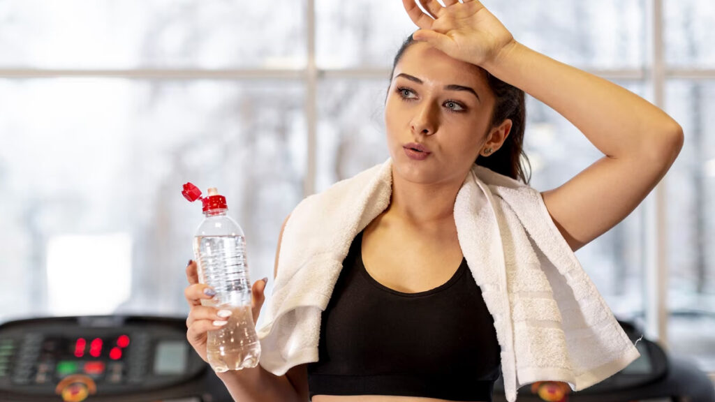 A young woman taking a break at the gym, holding an On Take Water bottle in one hand and a towel around her neck, showing the importance of hydration for physical performance and recovery.