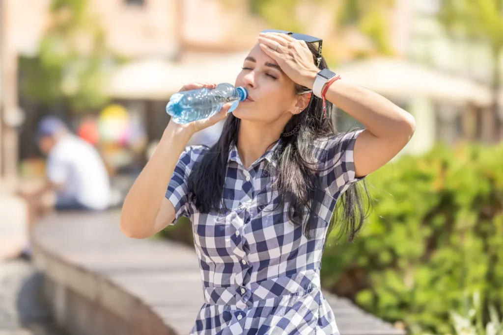 A woman cooling off under the sun while drinking from a water bottle, highlighting the importance of making On Take Water a daily habit to stay hydrated and energized throughout the day.