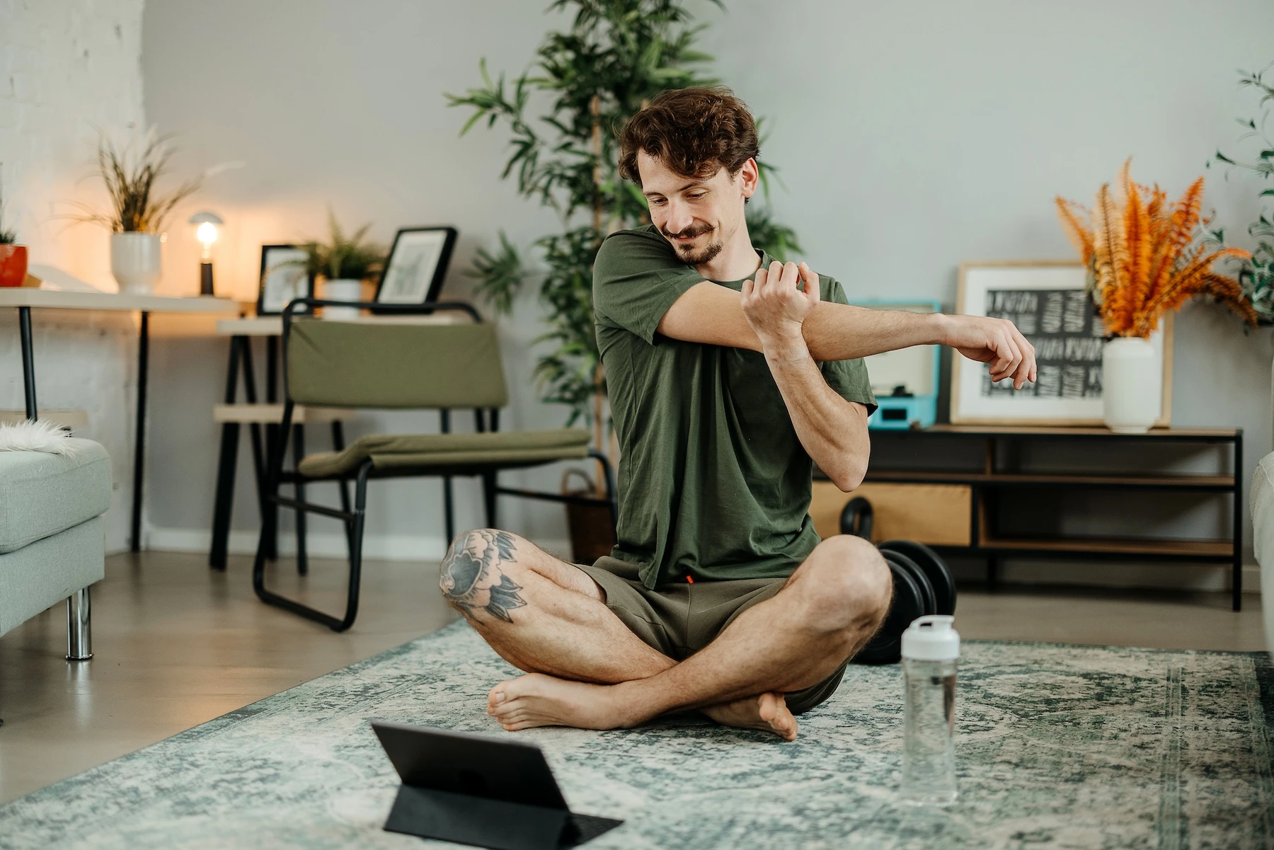 A man stretching indoors after a workout session, sitting cross-legged on a yoga mat with a water bottle labeled On Take Water nearby, emphasizing post-workout hydration.

