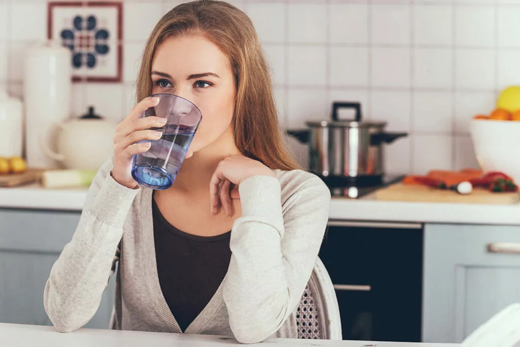 A young woman drinks a glass of clear water in a modern kitchen, representing the purity and health benefits of On-Take Water – naturally alkaline and mineral-rich.