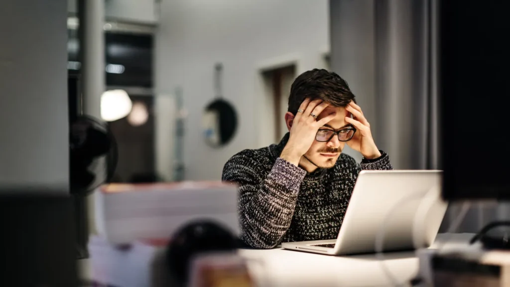 A man sitting at a desk with his hands on his forehead, looking stressed and unfocused while staring at a laptop screen, showing signs of brain fog and difficulty concentrating possibly due to dehydration and lack of water intake.