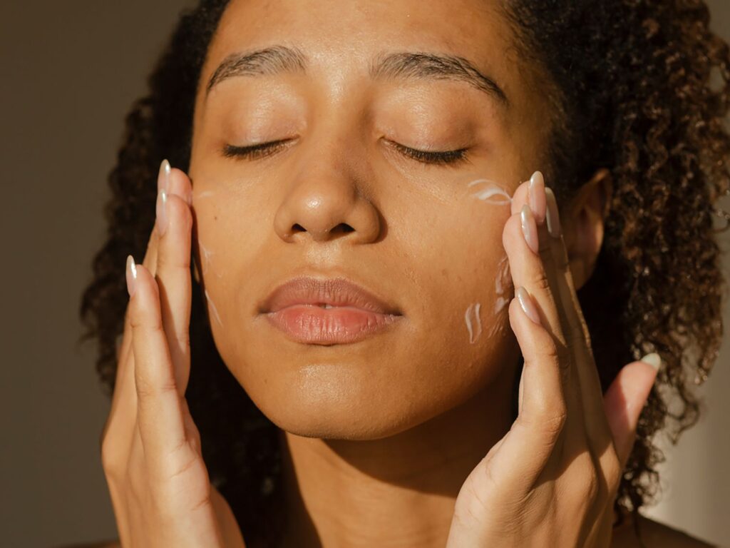A woman gently applying moisturizer to her face, showing signs of dry skin — a common result of dehydration. Drinking On Take Water daily helps improve skin hydration from within, reducing dryness and preventing breakouts naturally.