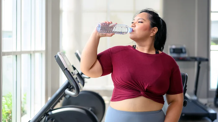 A woman in a maroon workout top and gray leggings is drinking water from a plastic bottle while standing near exercise equipment in a gym.