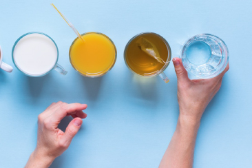 Top view of a person’s hands reaching for a glass of water among four different drinks, including milk, orange juice, and tea, on a light blue background. 