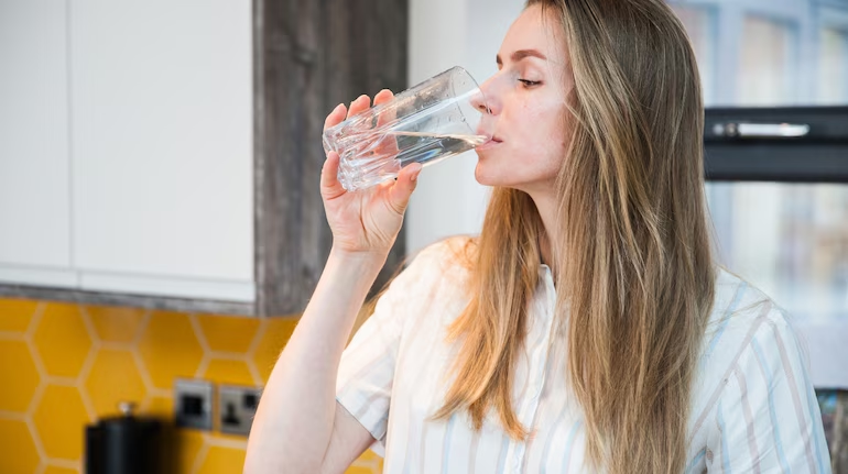 A young woman drinking a glass of water in a bright kitchen, illustrating the health benefits of starting your day with proper on take water.