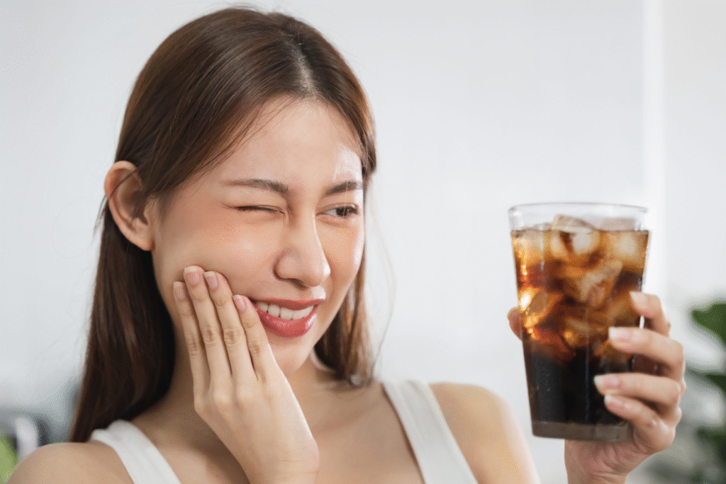 A young woman holding a glass of soda with ice while touching her cheek in discomfort, illustrating dental problems caused by sugary drinks.