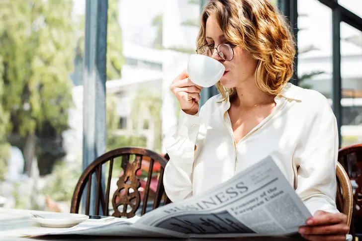 Woman in a white blouse reading a business newspaper and sipping coffee in the morning, representing a balanced energy strategy by combining water and caffeine wisely for focus and productivity.