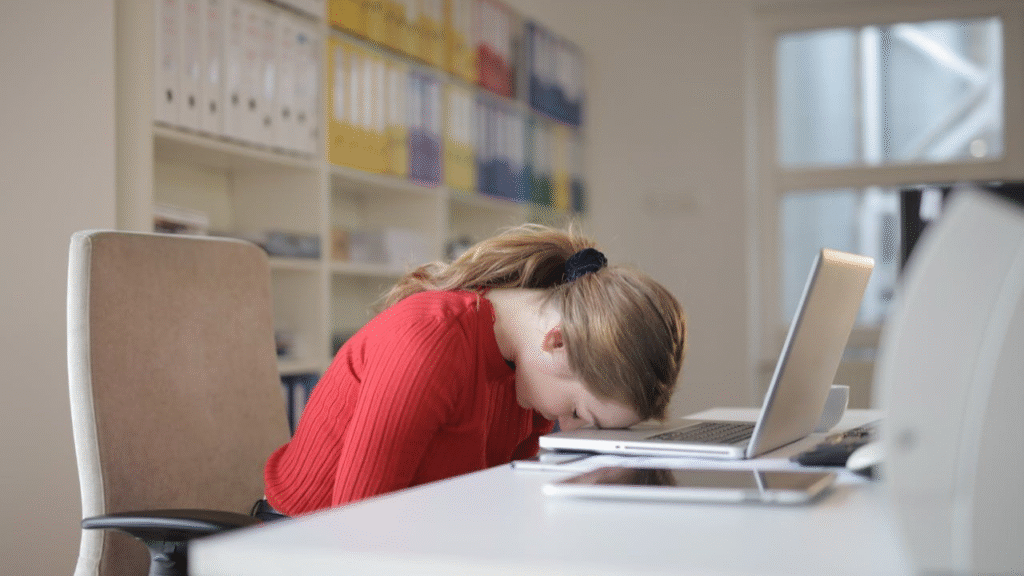 Woman in a red sweater with her head down on a desk beside a laptop, representing afternoon slump, fatigue, and the importance of managing energy with a balanced mix of water and caffeine.