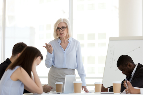 An older woman standing and speaking assertively during a meeting while team members appear frustrated and disengaged, representing mood swings and irritability that could be linked to dehydration or insufficient water intake.
